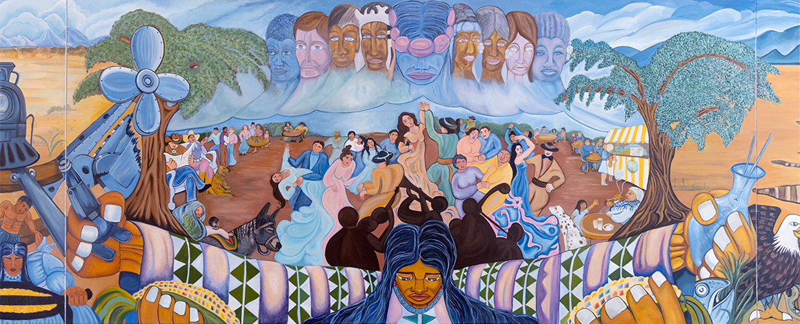 A vibrant mural depicting a gathering of people beneath a large cloud of faces, surrounded by trees, mountains, a train, and an eagle. The scene includes musicians, dancers, and colorful traditional patterns.