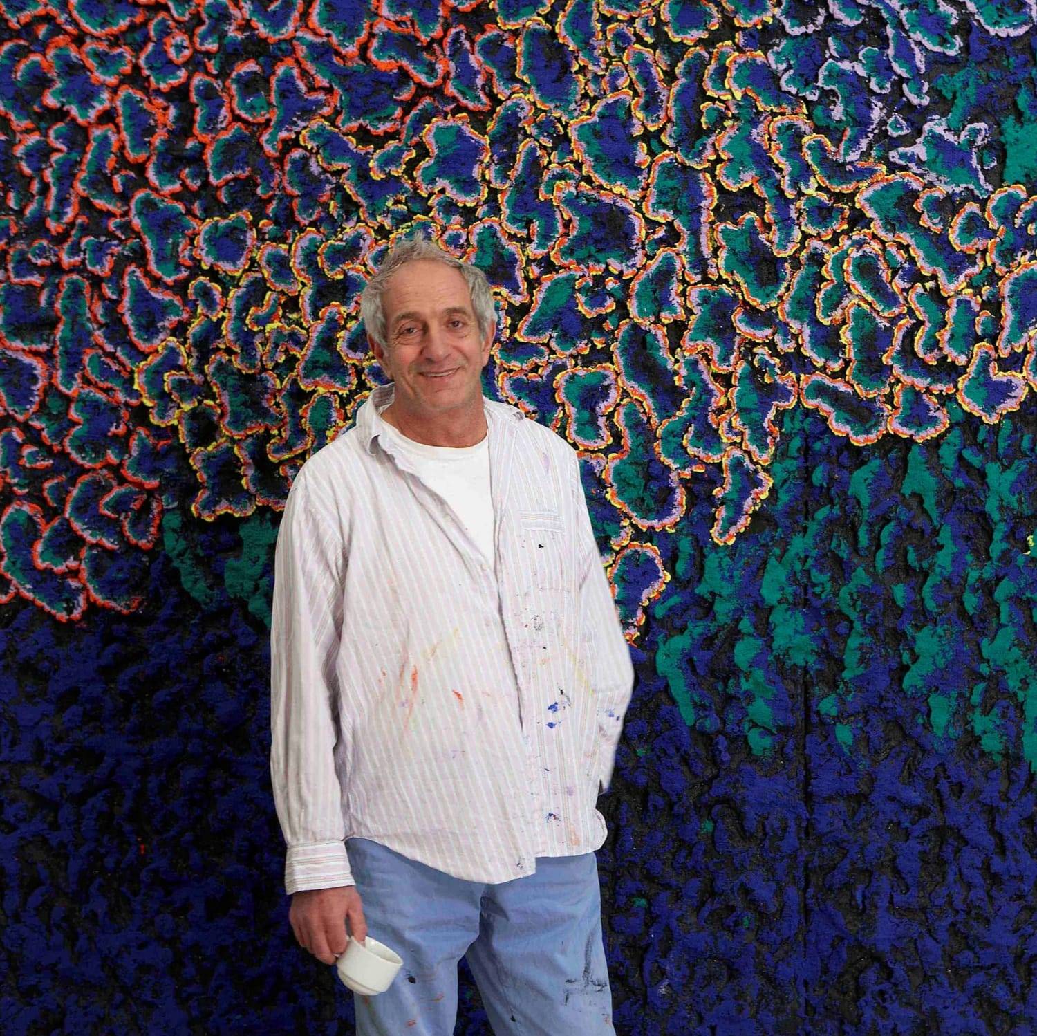 A person wearing a paint-splattered shirt and jeans stands in front of a vibrant abstract artwork featuring intricate, colorful patterns of orange, blue, green, and purple.